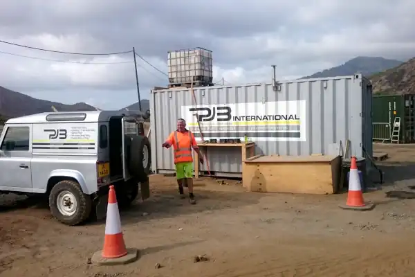 A PJB International mobile laboratory container on a remote construction site, with a PJB-branded Land Rover Defender and a worker in high-visibility clothing, set against a mountainous backdrop.