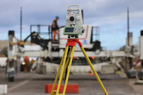 A Leica total station on a yellow tripod in the foreground, with a construction worker and paving machinery visible on site in the background.